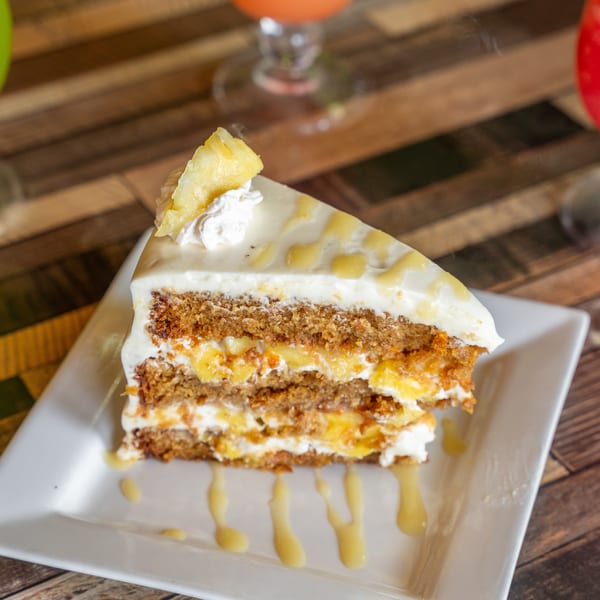 A slice of pineapple carrot cake with cream cheese frosting and pineapple chunks on a white plate.