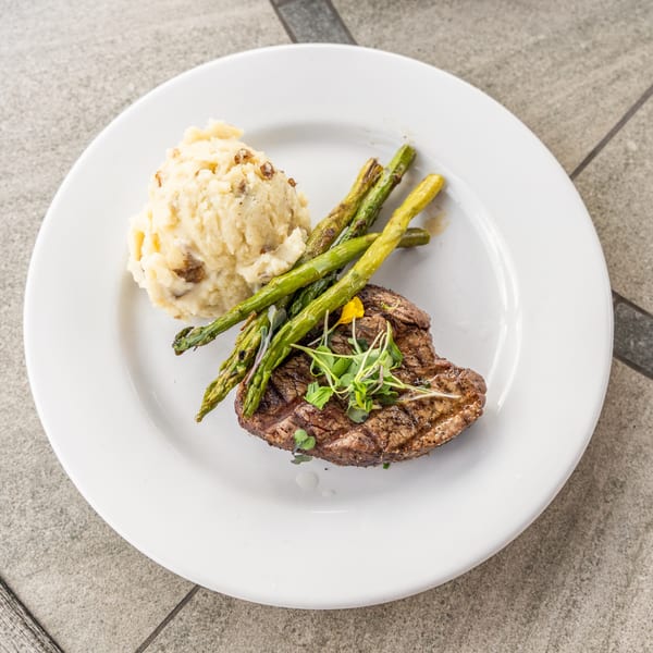 Grilled steak with mashed potatoes and asparagus, garnished with microgreens and a yellow flower.