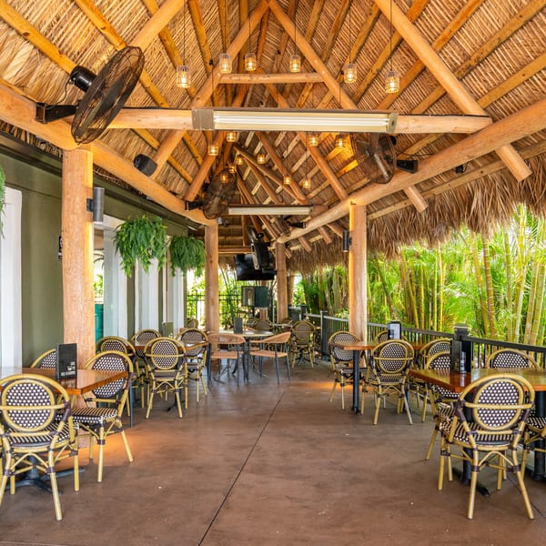 Outdoor restaurant patio with a thatched roof, wooden tables, and woven chairs, surrounded by lush greenery.