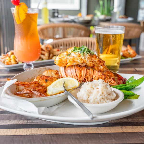 Grilled lobster tail, shrimp, rice, and snap peas with a cocktail and beer on a wooden table.
