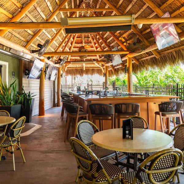 An outdoor bar and dining area with a thatched roof, bar stools, and round tables.