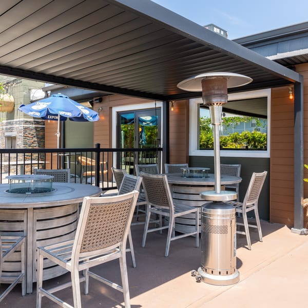 Outdoor patio with wooden tables, chairs, and a blue umbrella under a black awning on a sunny day.
