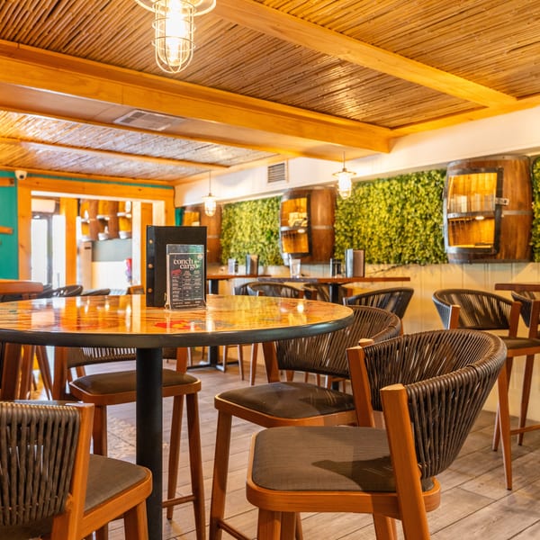 Restaurant interior with high-top tables, woven chairs, bamboo ceiling, and a green wall with barrel decor.