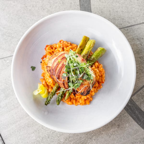 Bacon-wrapped chicken with microgreens, asparagus, and tomato risotto in a white bowl.