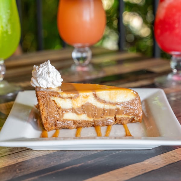 Slice of caramel cheesecake with whipped cream on a white plate, with colorful drinks in the background.