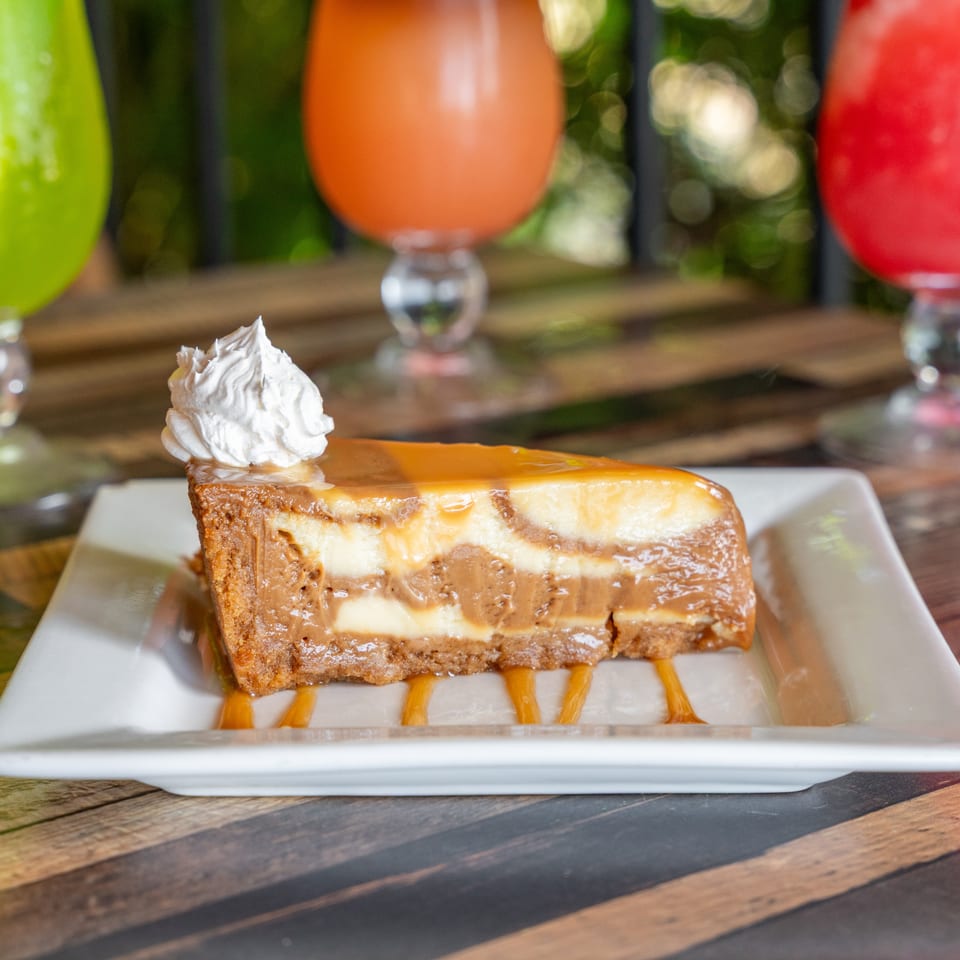 Slice of caramel cheesecake with whipped cream on a white plate, with colorful drinks in the background.