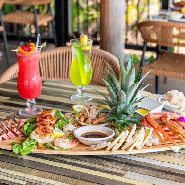 A large seafood platter with crab legs, shrimp, tuna, and calamari, alongside two colorful cocktails.