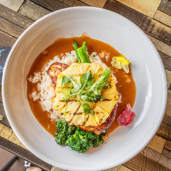 Grilled pineapple and chicken over rice with sauce, broccolini, and microgreens in a white bowl.