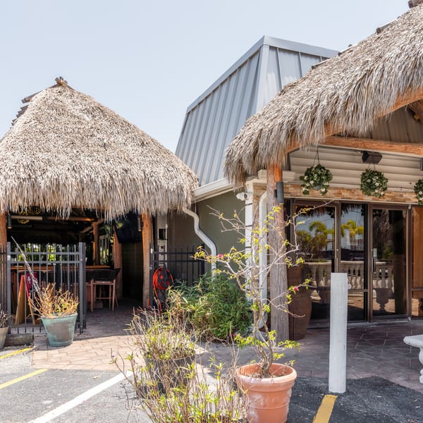 Exterior of a restaurant with two tiki huts, a modern building, and potted plants.