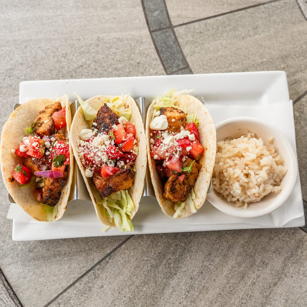 Three fish tacos with salsa, cheese, and a side of rice on a white rectangular plate.
