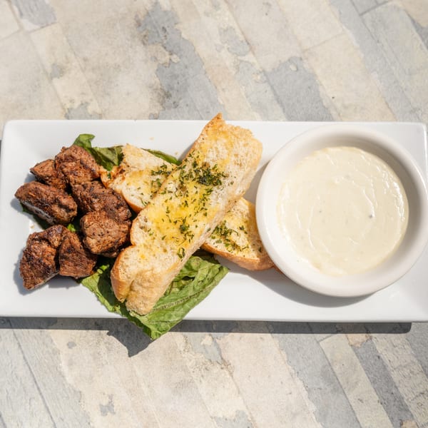 Grilled steak bites, garlic bread, and a side of creamy white sauce on a rectangular plate.