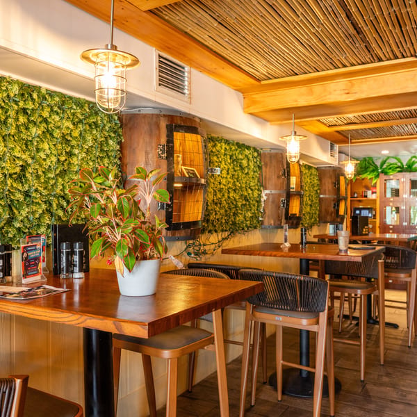 Restaurant interior with wooden high-top tables, woven chairs, green wall decor, and barrel accents.