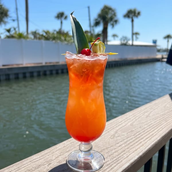 Tropical orange cocktail with pineapple leaf, cherry, and pineapple wedge on a dock railing overlooking water.