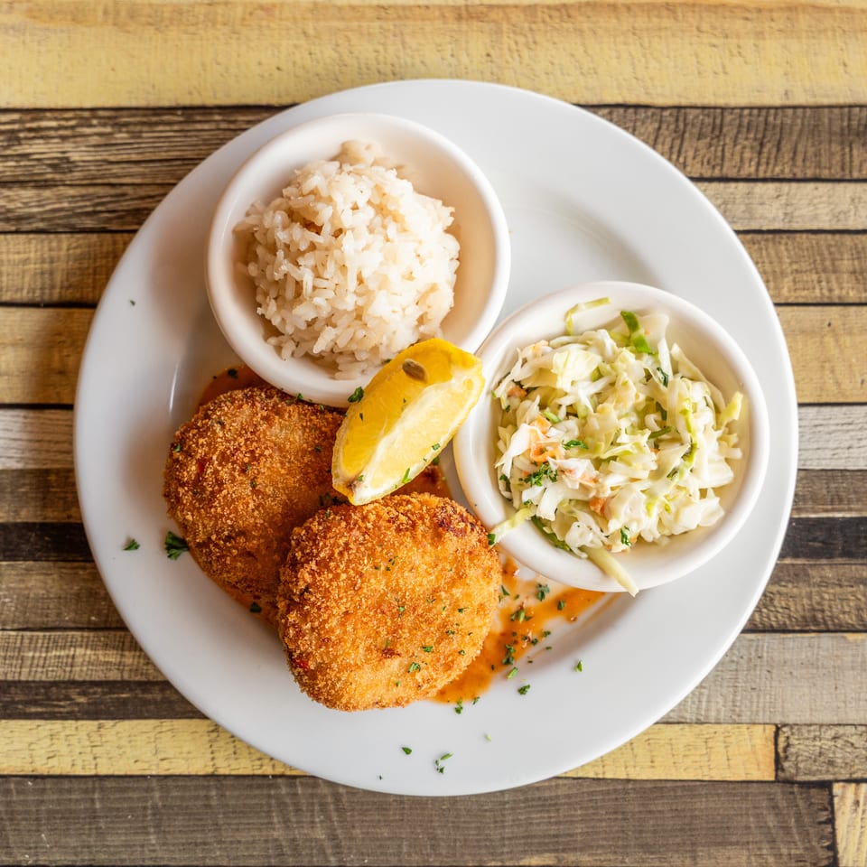 Two golden-brown crab cakes with rice, coleslaw, and a lemon wedge on a white plate.