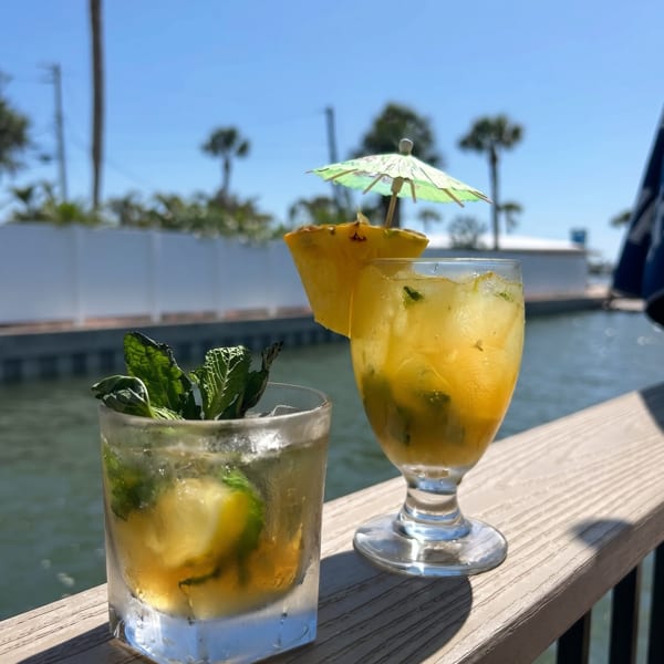 Two tropical drinks with mint and pineapple on a wooden railing overlooking a canal with palm trees.