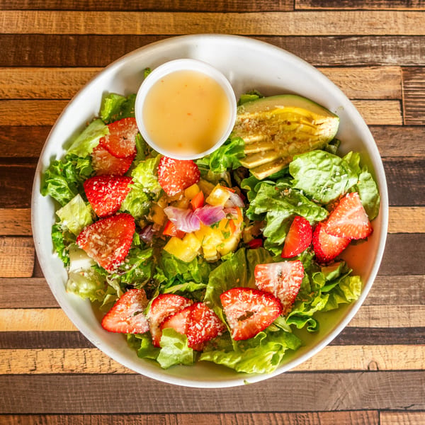 Fresh green salad with sliced strawberries, avocado, pineapple salsa, and dressing in a white bowl.