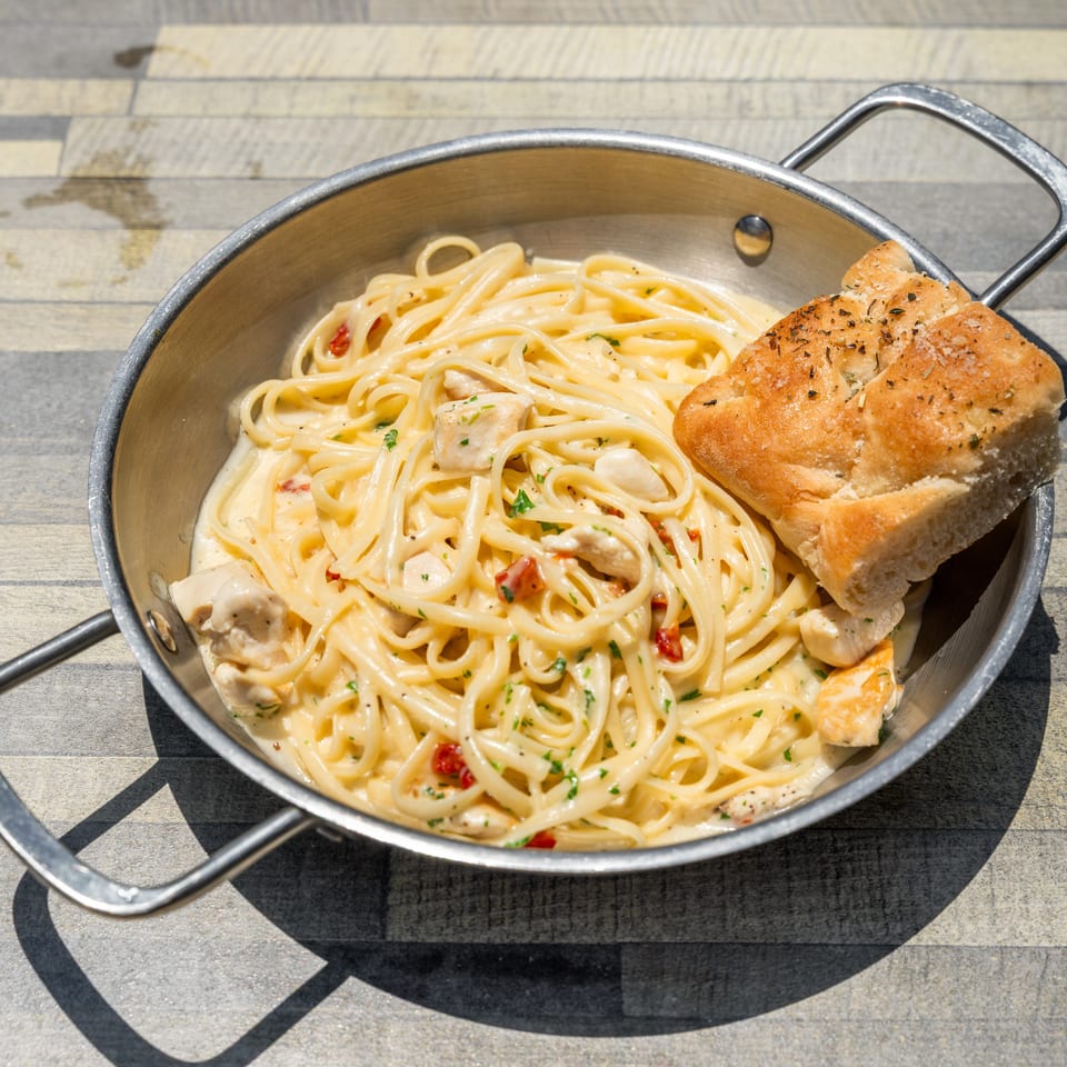Creamy chicken linguine pasta with sun-dried tomatoes and a side of focaccia bread in a metal pan.